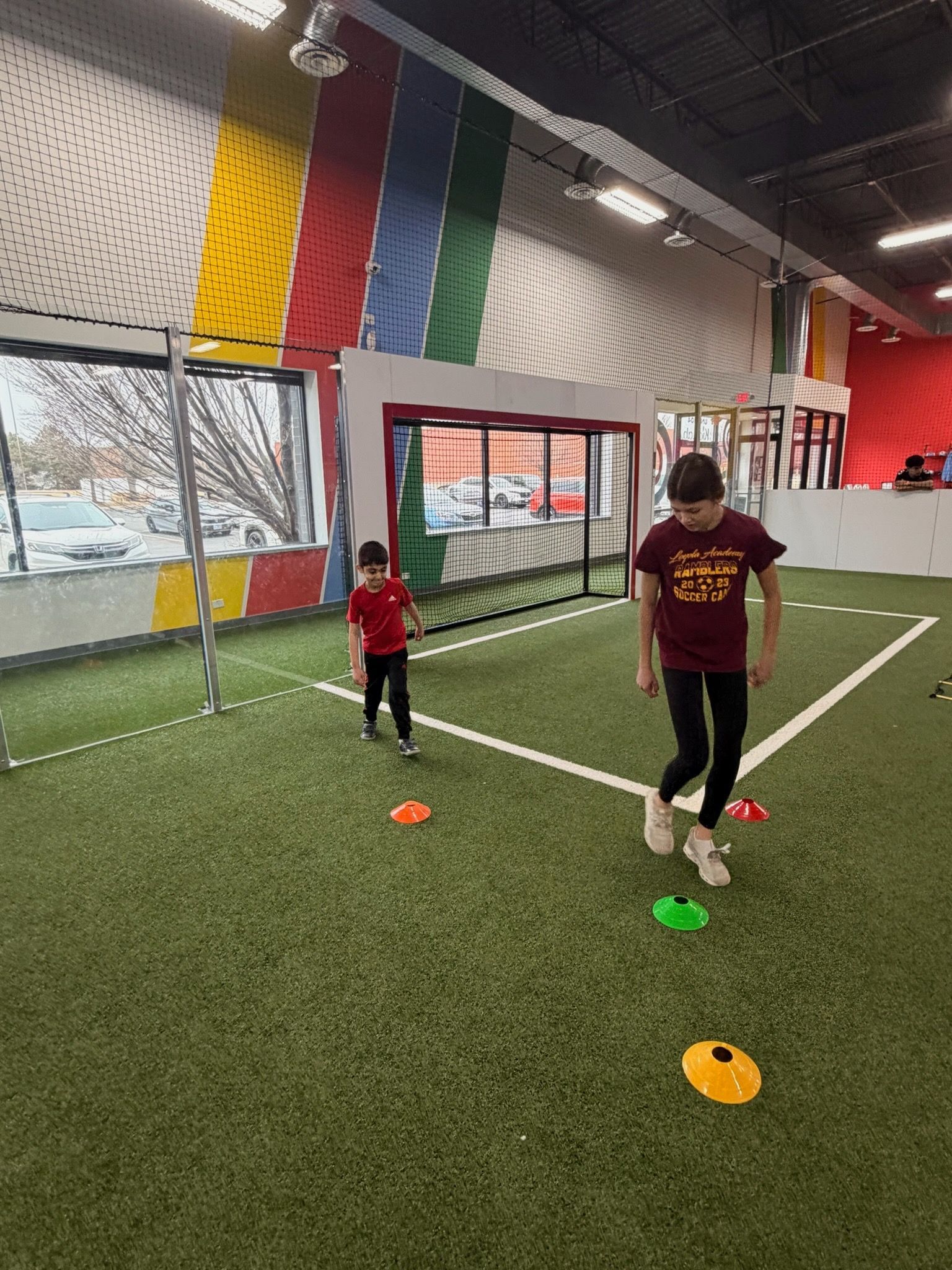 Two people training on indoor turf with colored cones and a goal net in the background