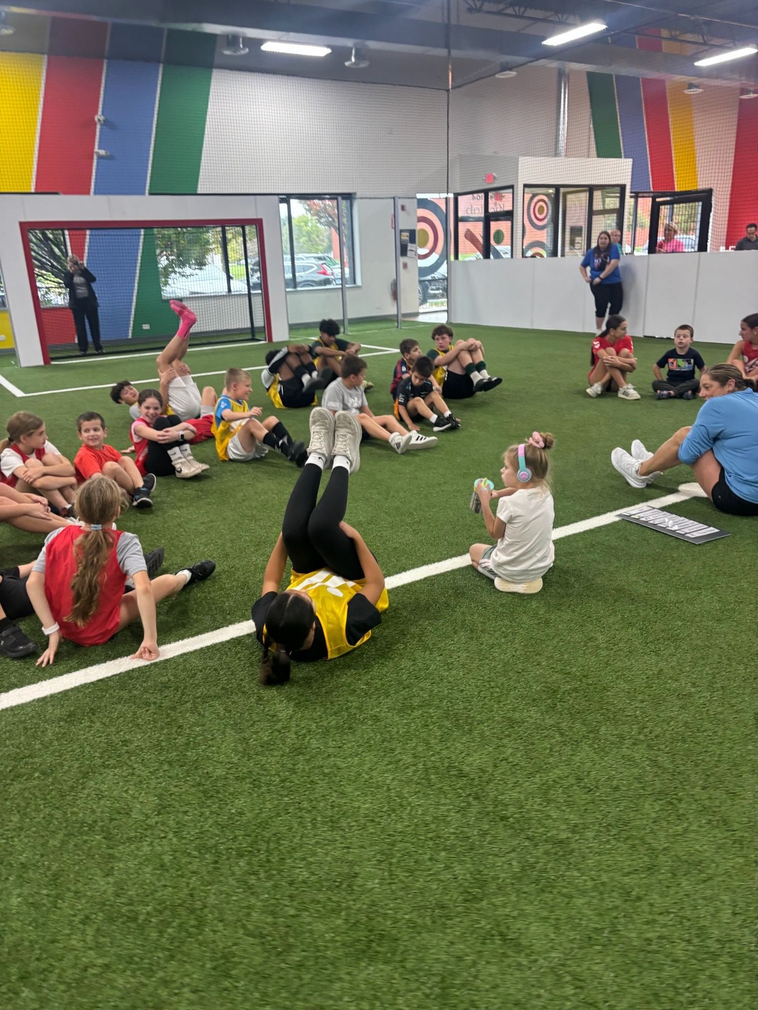 Children stretching on turf inside an indoor sports facility with colorful wall panels and a large mirror.