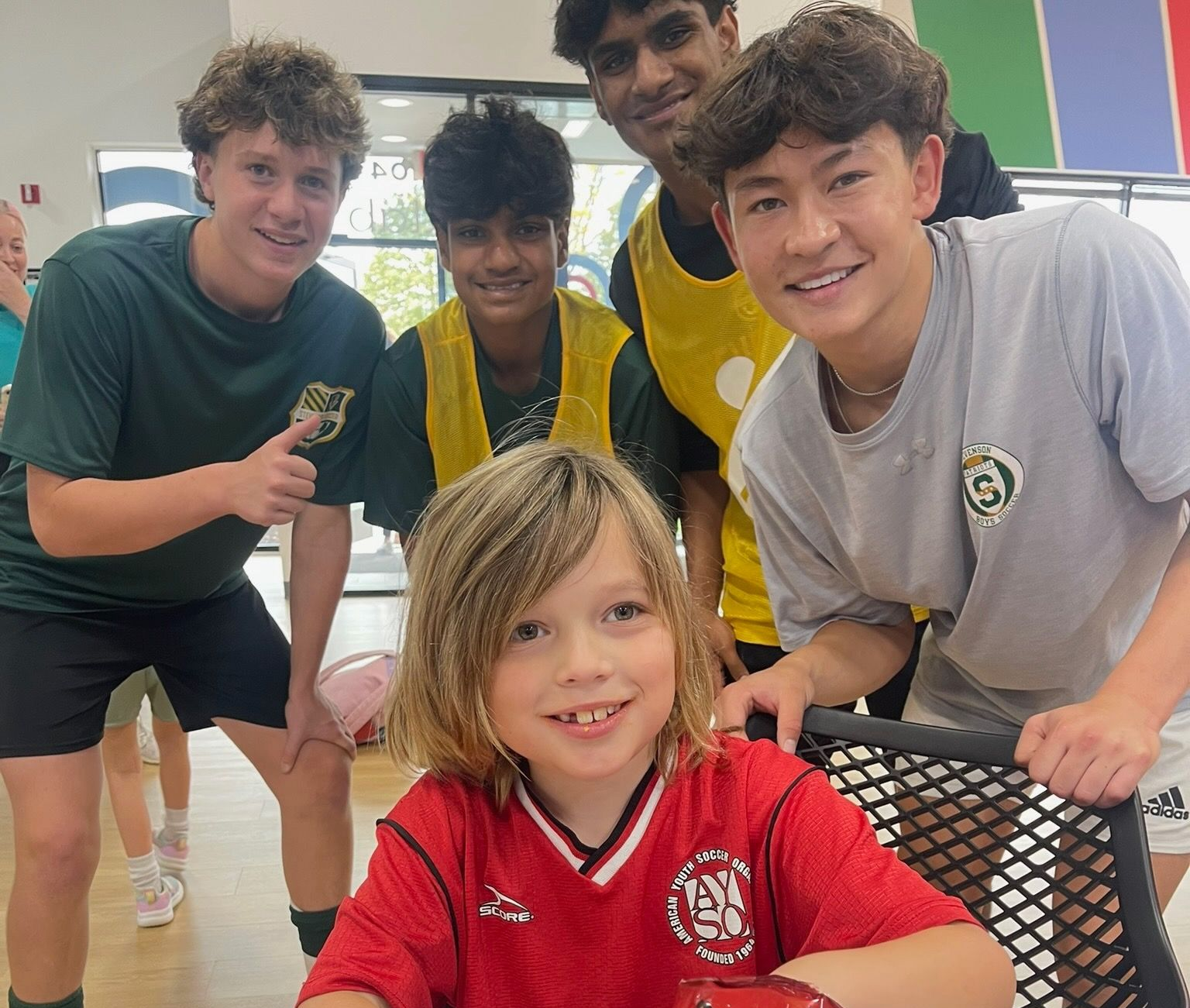 Five smiling kids pose in a gym, leaning toward the camera around a seated child in a red shirt.
