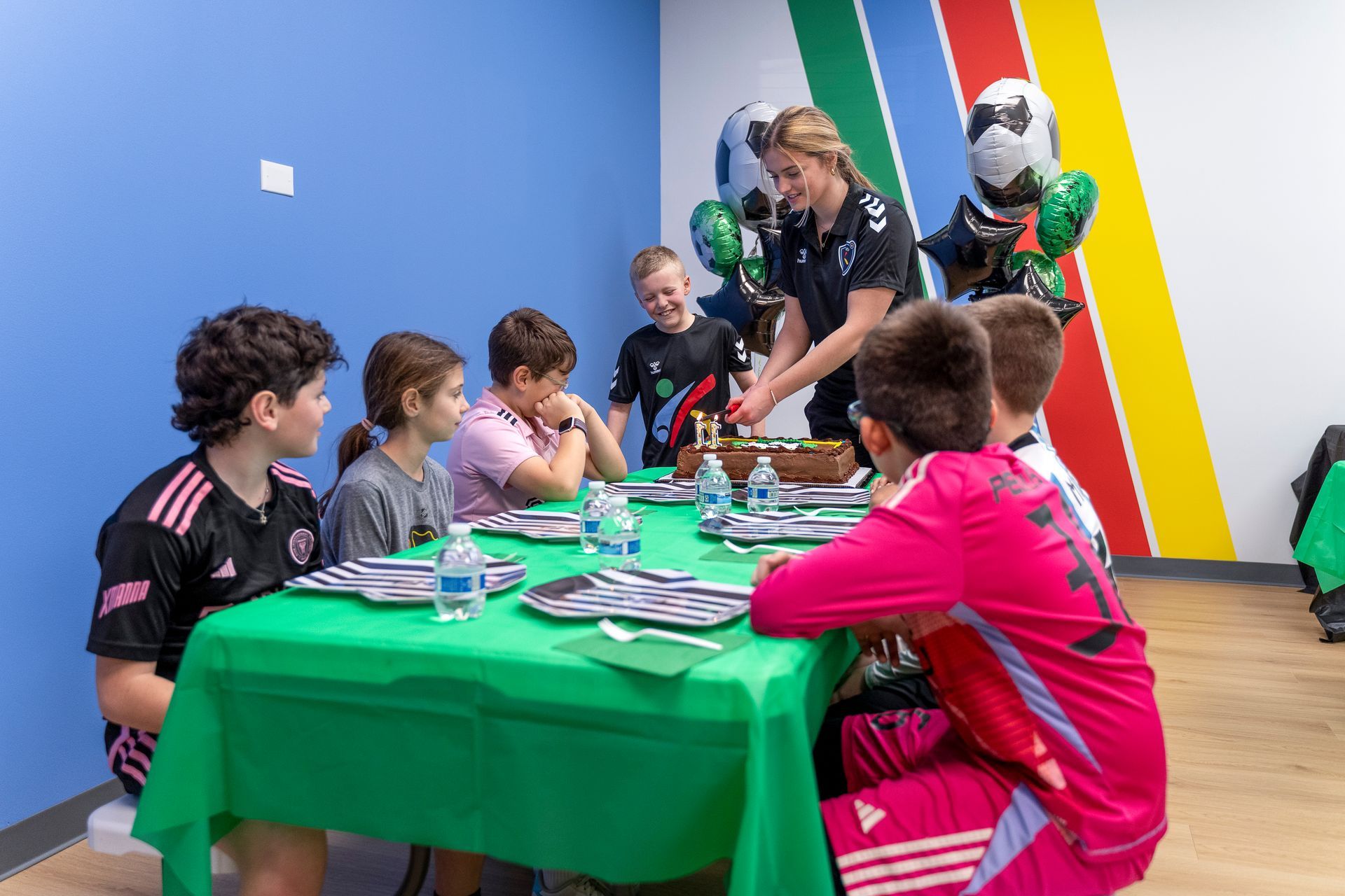 Chess players gathered around a table in a colorful room, with two standing spectators leaning over the board.