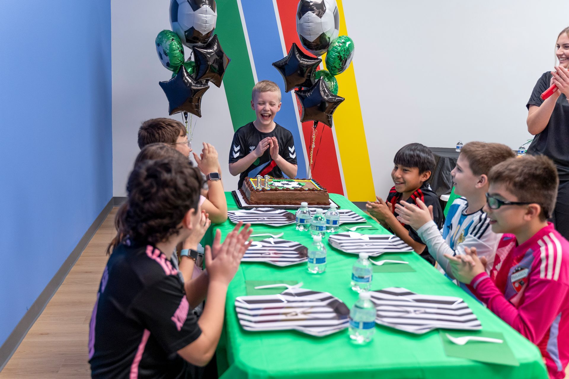 Children gathered around a green table with craft supplies in a bright classroom, smiling and clapping.