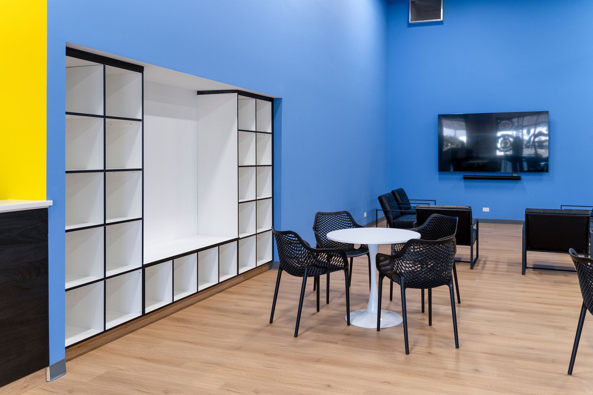 Modern lounge with blue walls, white shelving, round table, black chairs, and a wall-mounted TV
