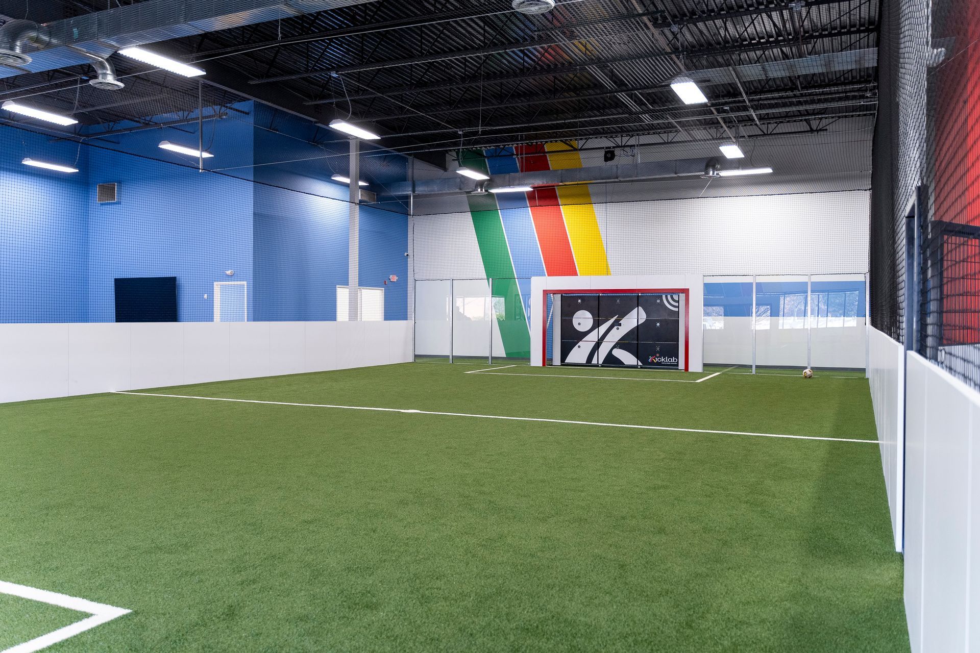 Indoor soccer field with green turf, white boards, and a goal against a wall with colorful vertical stripes