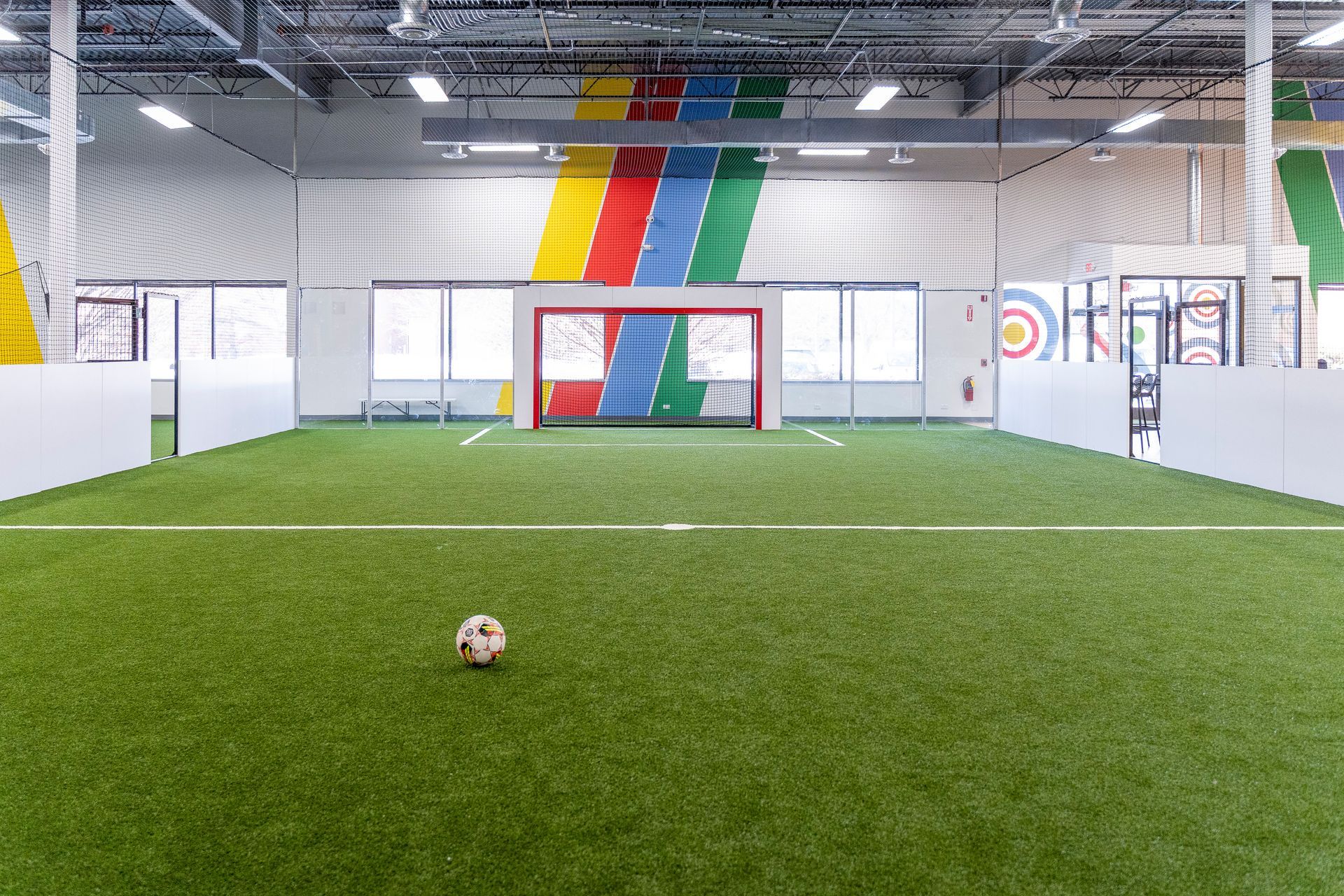 Indoor soccer field with a lone soccer ball on green turf and a red goal against a wall with colorful stripes.