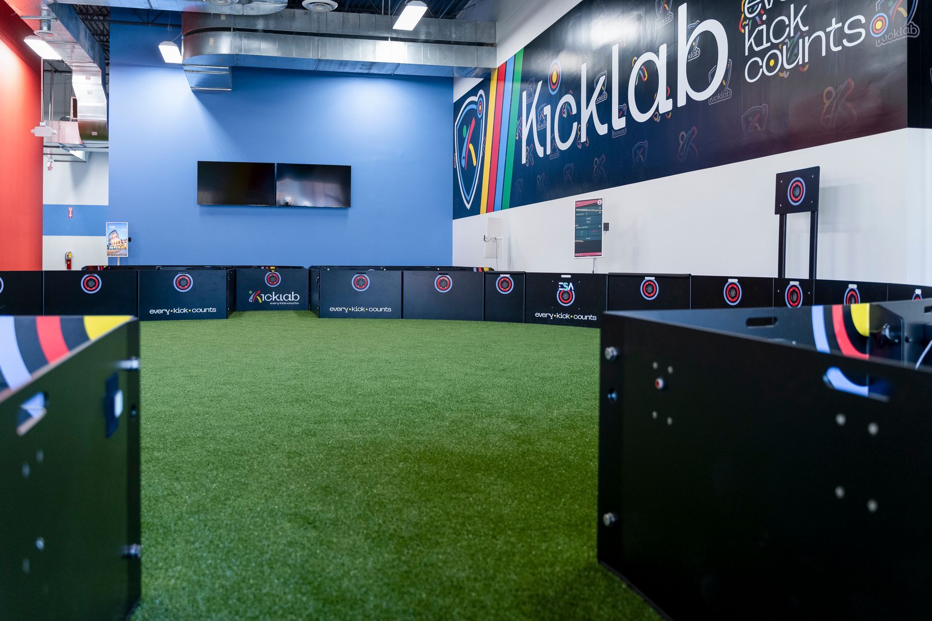 Indoor turf sports court with black barriers and a large wall graphic reading “The Lab Back About Us”