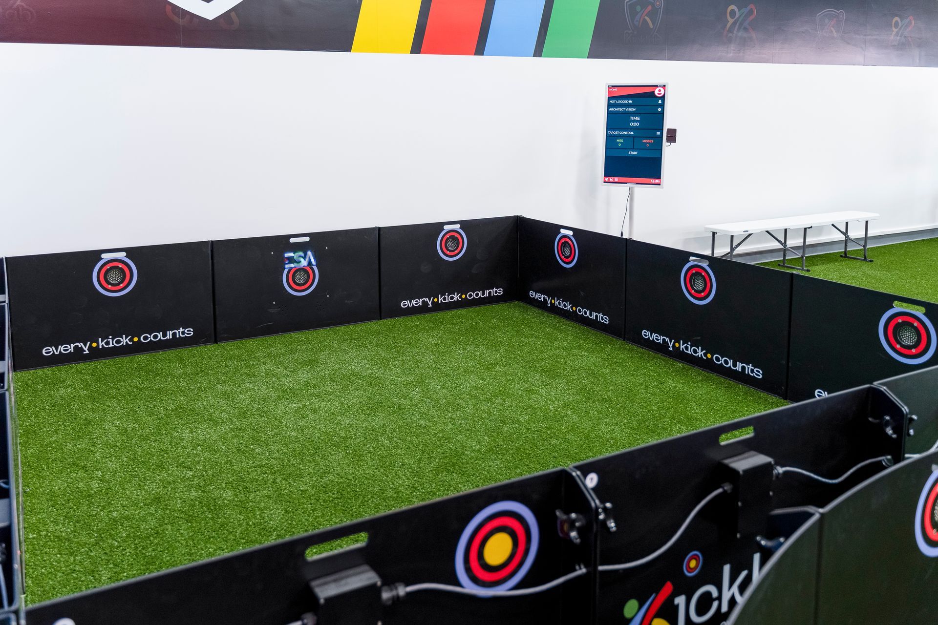 Small indoor mini-soccer arena with black padded walls and artificial turf beside a white wall.