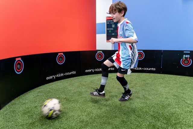 Child playing soccer in a red-and-black indoor arena, kicking a ball on artificial turf.