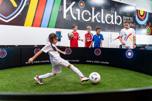 Child kicking a soccer ball in a Kicklab play area with spectators behind a barrier