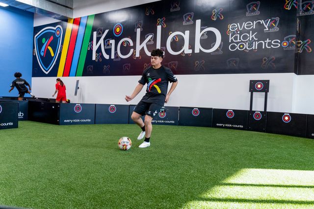 Soccer player in black kicks a ball on indoor turf at a Kicklab facility
