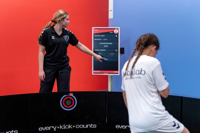 Two people at a sports event, one pointing at a scoreboard on a red and blue court