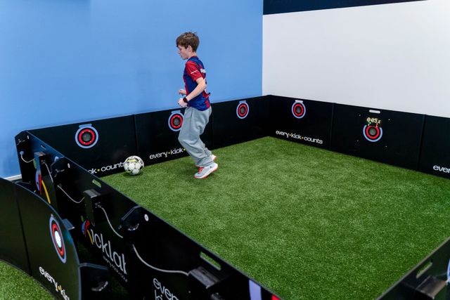Child in a soccer practice area with black barriers, chasing a ball on artificial turf.