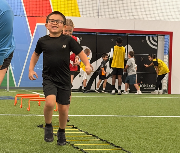 Child running an agility ladder on indoor turf, with soccer goal and other children in the background