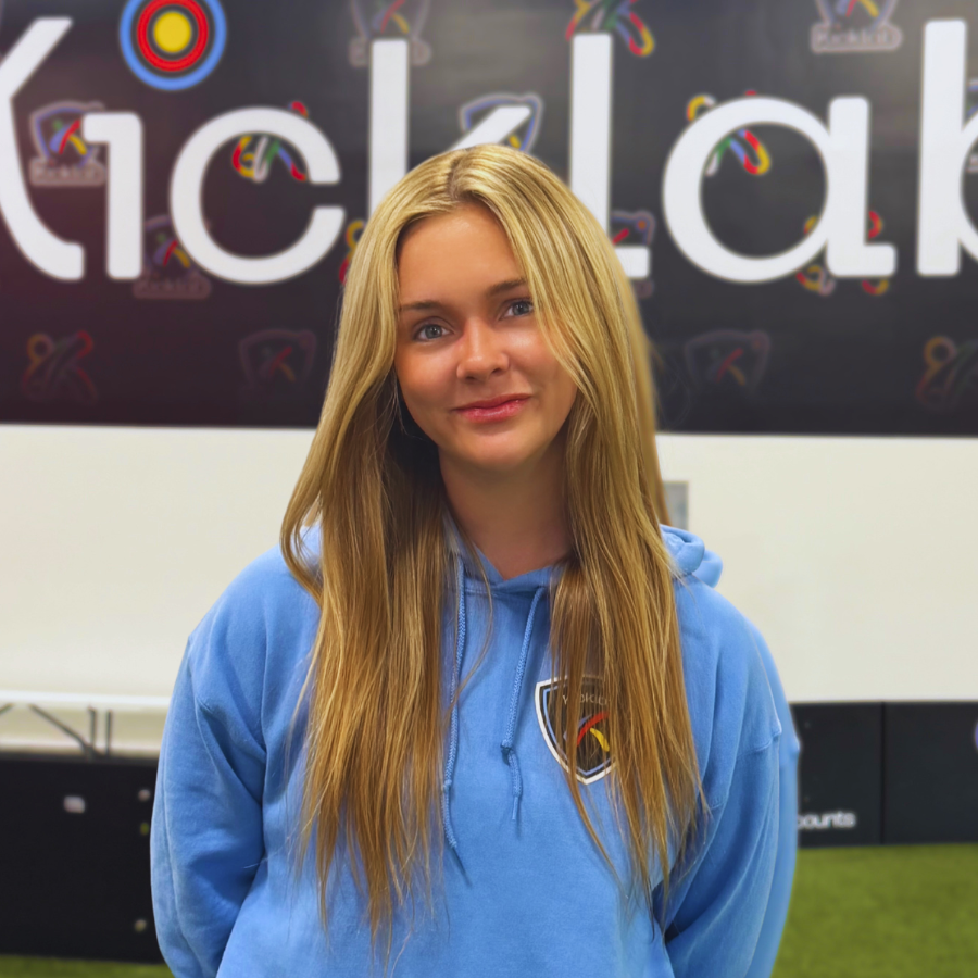 Young person in a blue hoodie standing indoors in front of a colorful sign, smiling at the camera