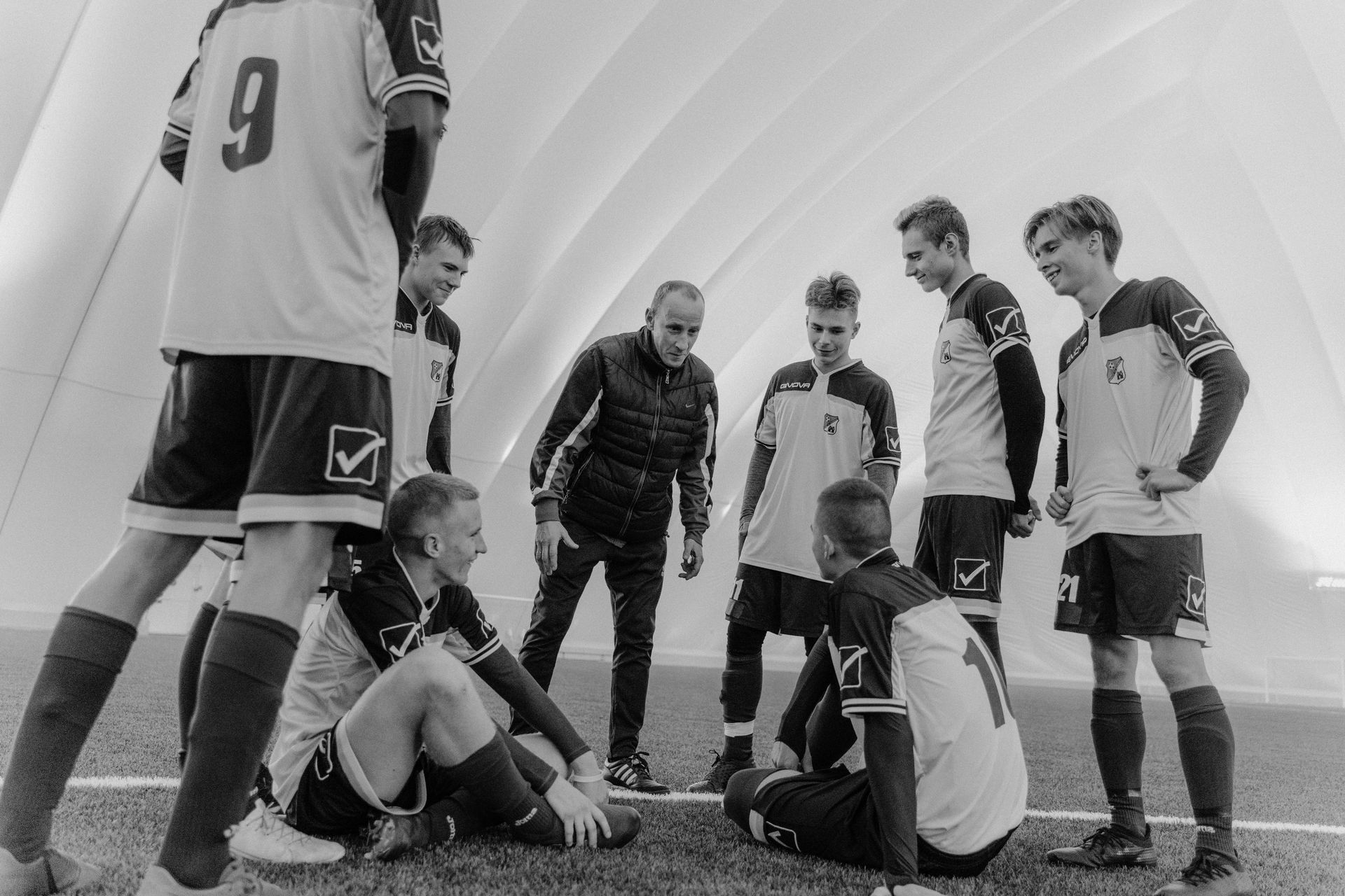 Soccer players huddle on the field during a break, with one seated on the grass.