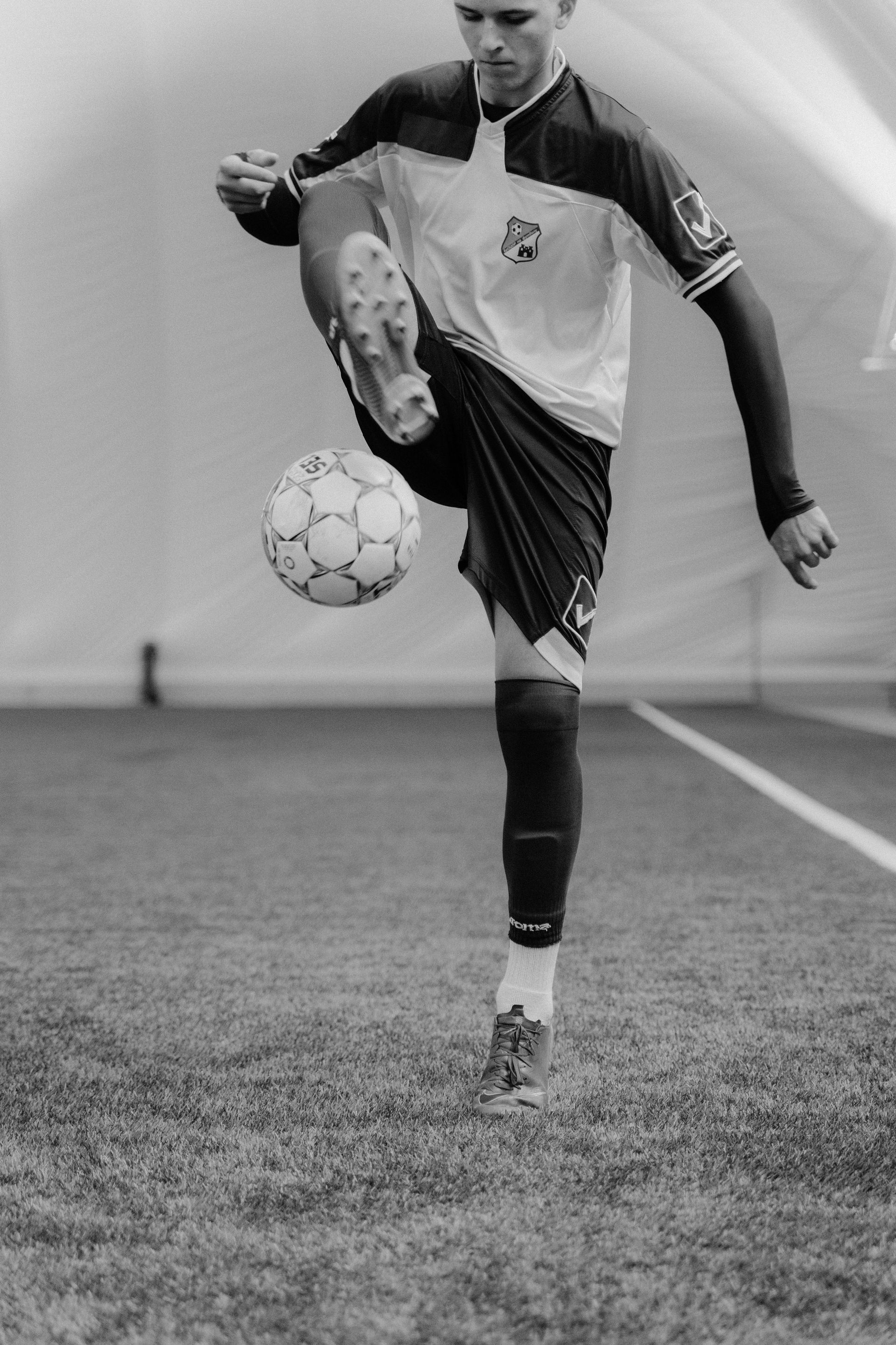 Soccer player juggling a ball on an indoor field, mid-kick in black and white.
