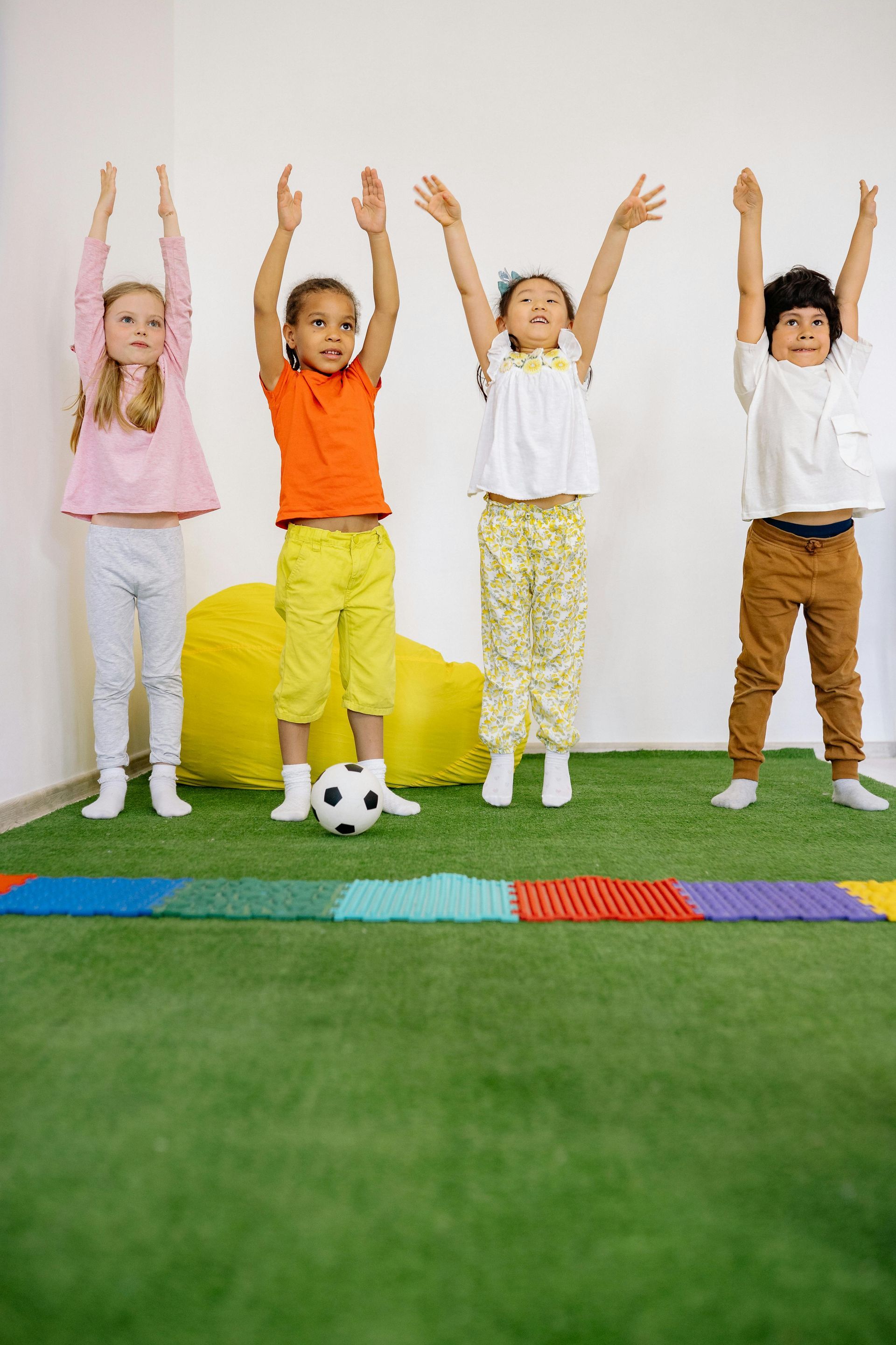 Four children standing with arms raised in a bright room, on colorful floor mats and grass-like carpet