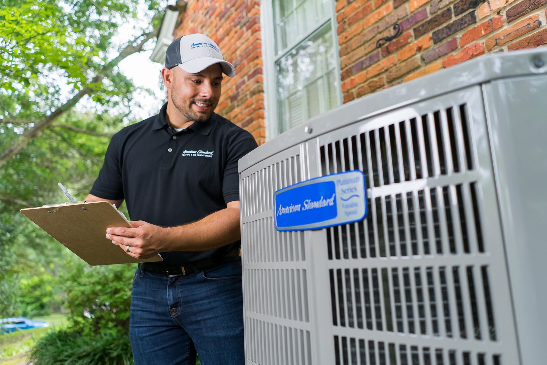 HVAC technician inspecting an American Standard AC unit outdoors. He holds a clipboard, smiling.