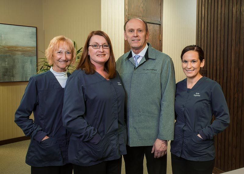 Four people in dark uniforms and a green jacket standing together indoors, smiling at the camera. Four people in dark uniforms and a green jacket standing together indoors, smiling at the camera.
