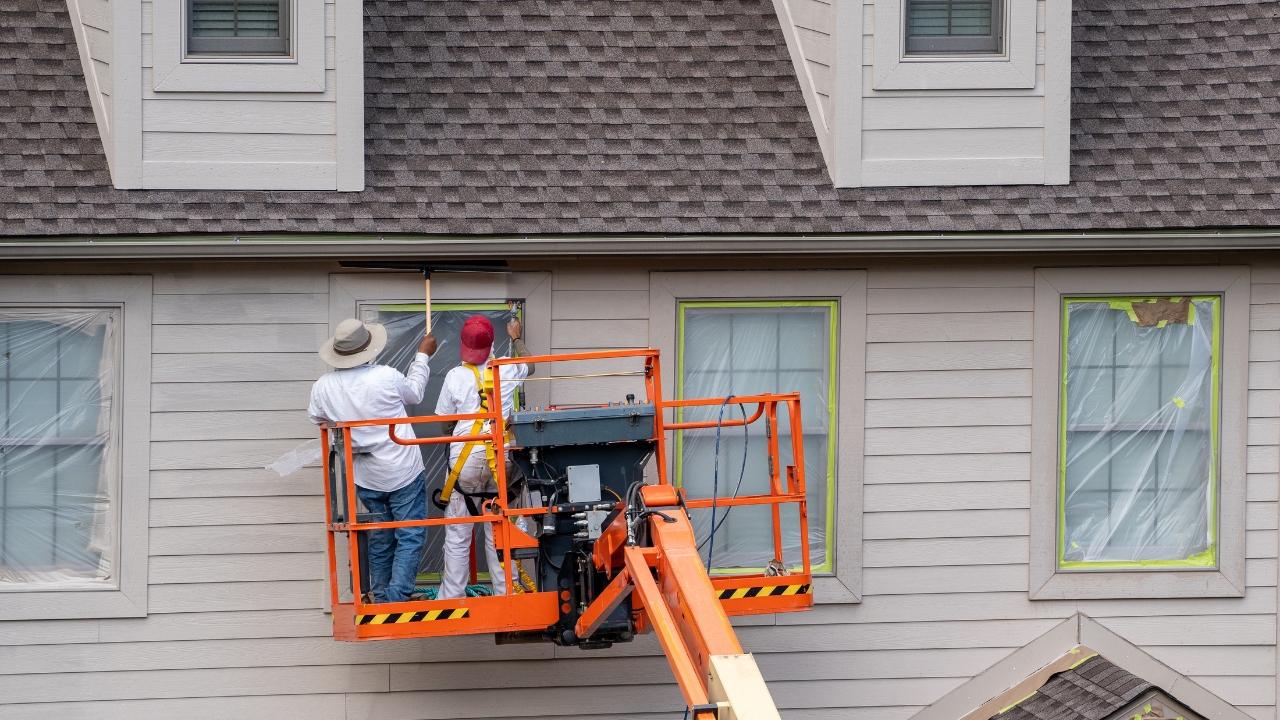Two people in a bright orange aerial lift painting the exterior of a light-colored two-story house.