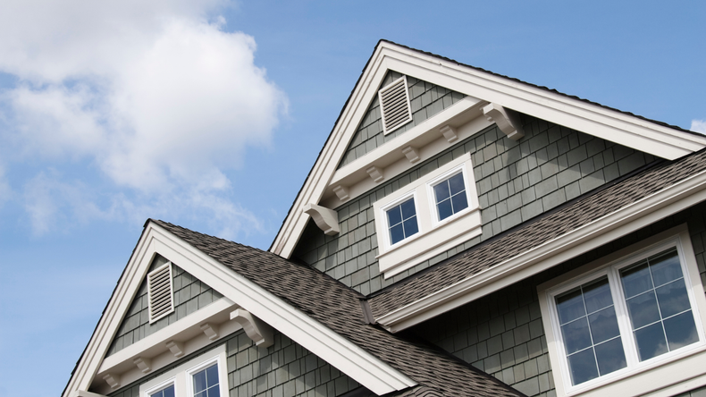 The peaked gables of a sage green, shingled home under a blue sky with white clouds.