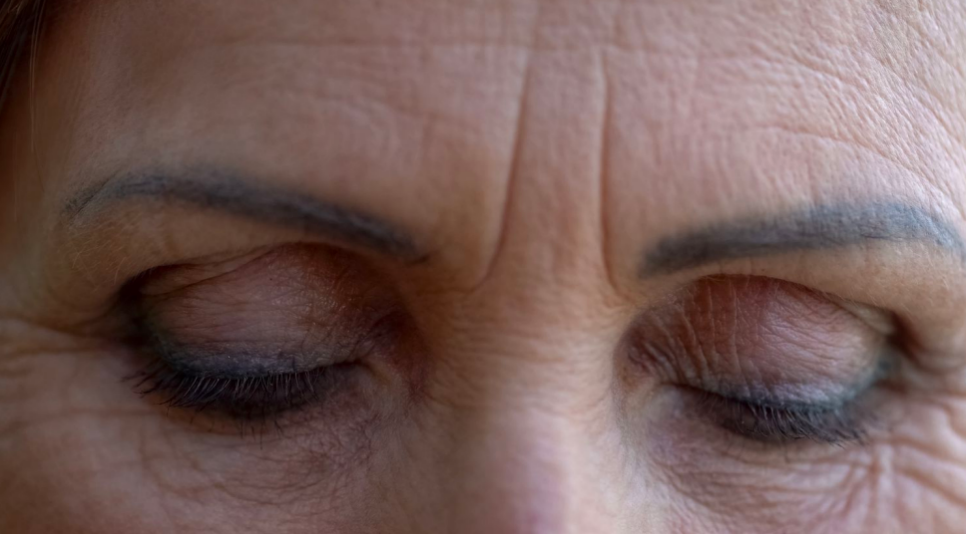 Woman's eyes closed, surrounded by wrinkles, dark eyebrows, and subtle makeup.
