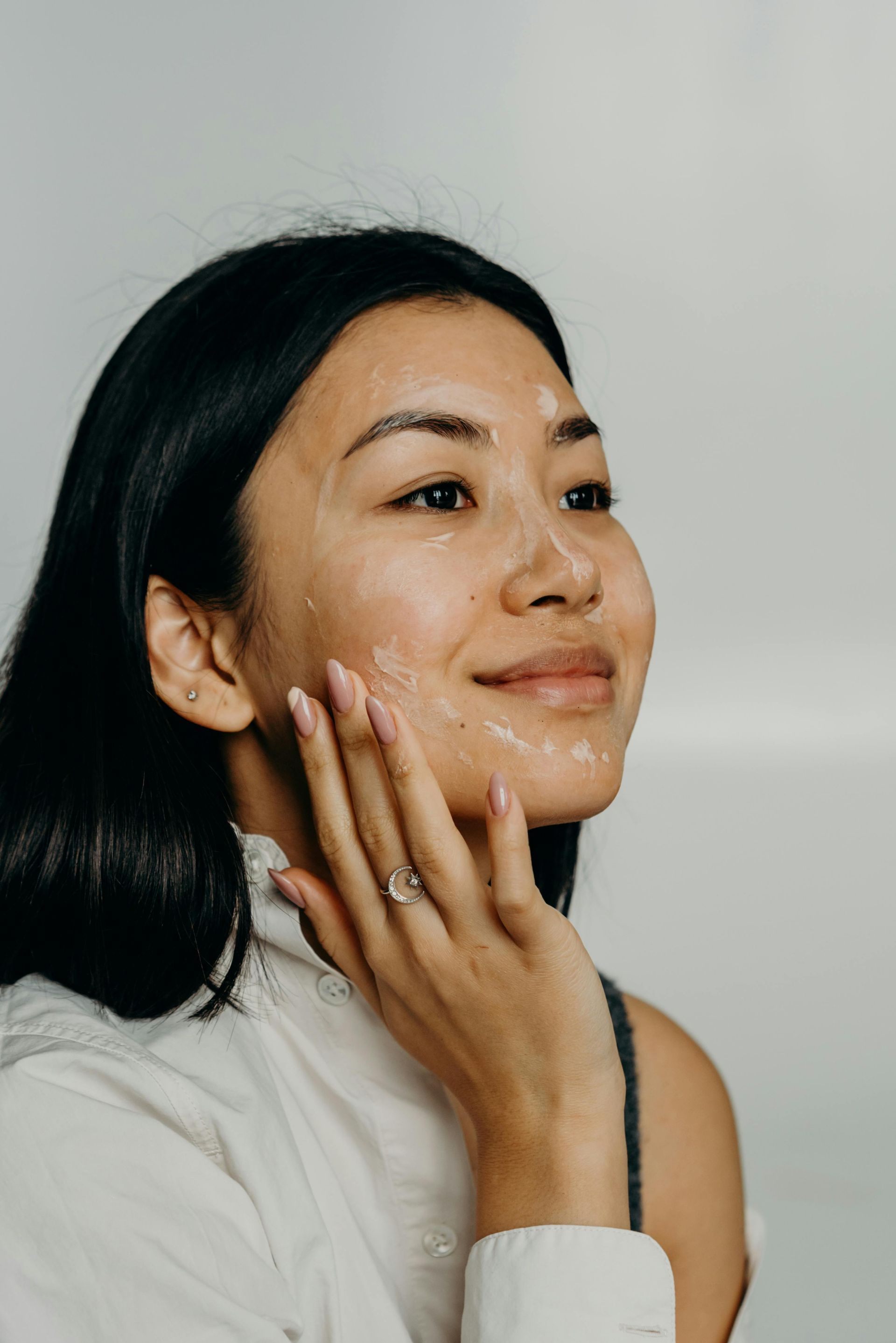 Woman with black hair applying face mask, smiling, touching her cheek.