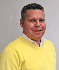 A man wearing a yellow sweater and a white shirt is smiling for the camera.