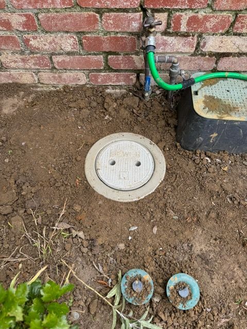 A manhole cover is sitting in the dirt next to a brick wall.