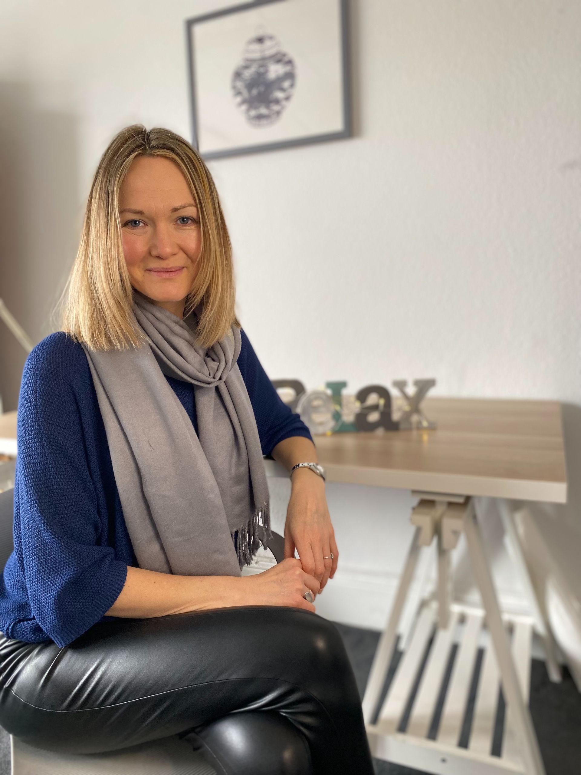 Woman seated on a chair in a modern office, smiling beside a desk and framed wall art.
