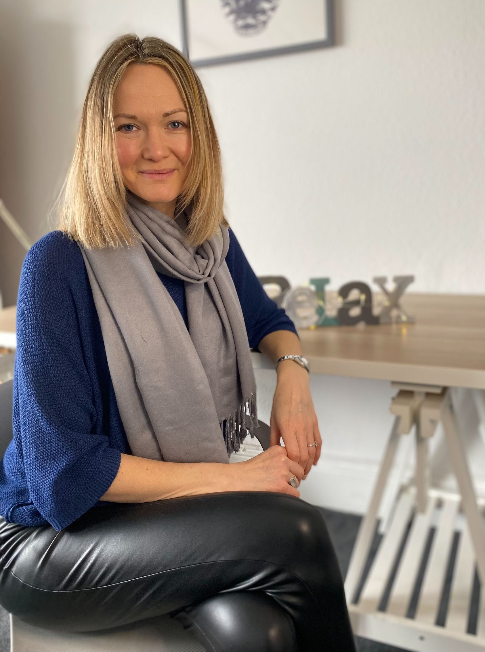 Woman seated indoors in a blue sweater and gray scarf, smiling beside a table.