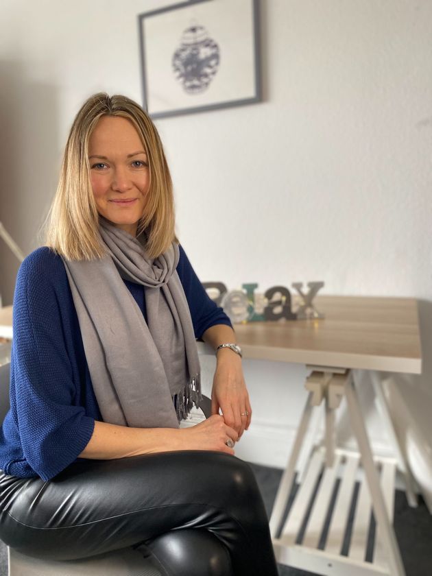 Woman seated by a desk in a bright room, wearing a blue sweater and gray scarf, smiling at the camera.