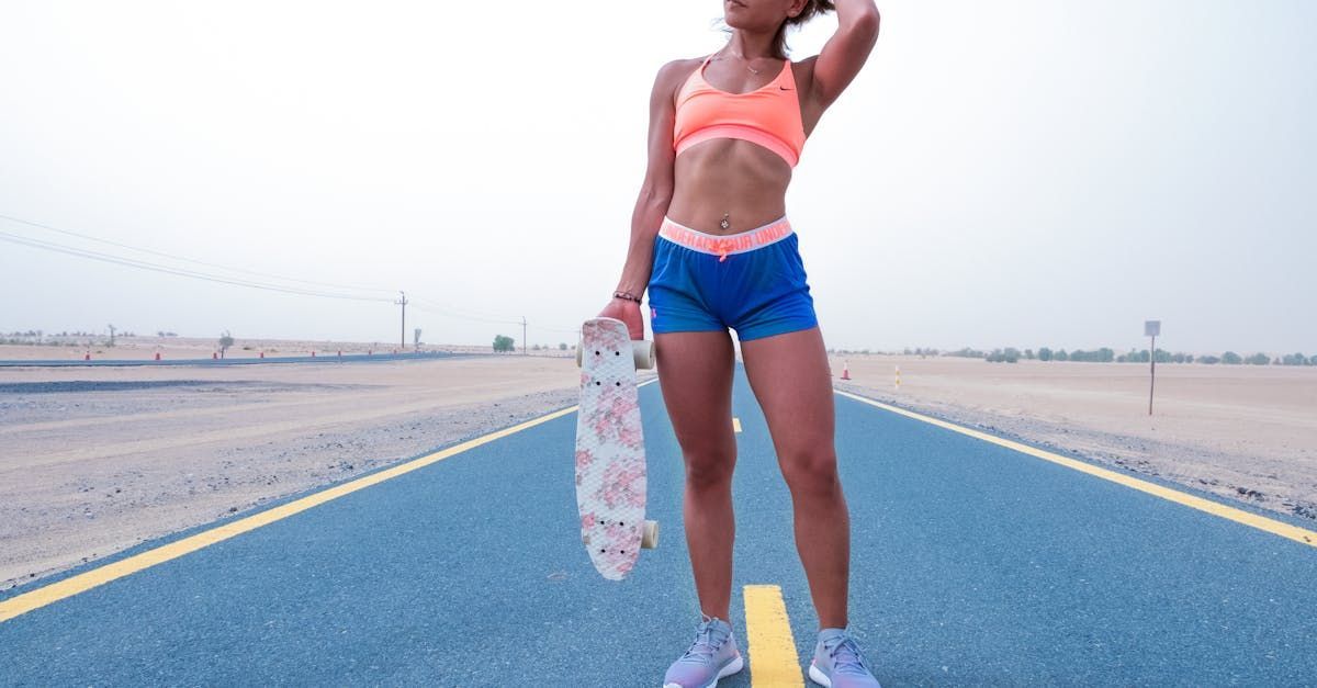 Person standing on an empty road holding a skateboard, wearing a pink top and blue shorts.
