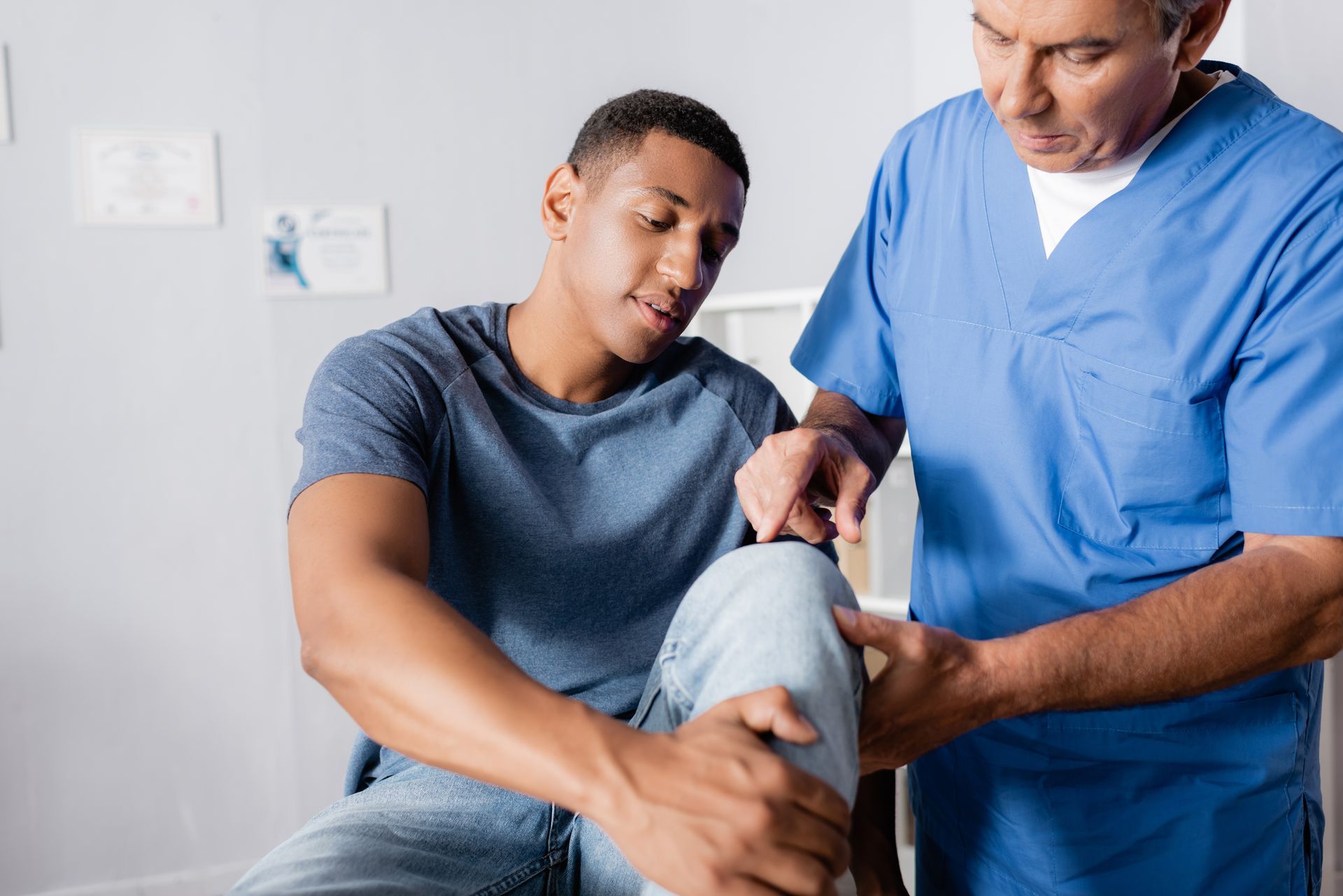 Doctor in blue scrubs examines a seated male patient's painful, injured knee during an evaluation. Doctor in blue scrubs examines a seated male patient's painful, injured knee during an evaluation.
