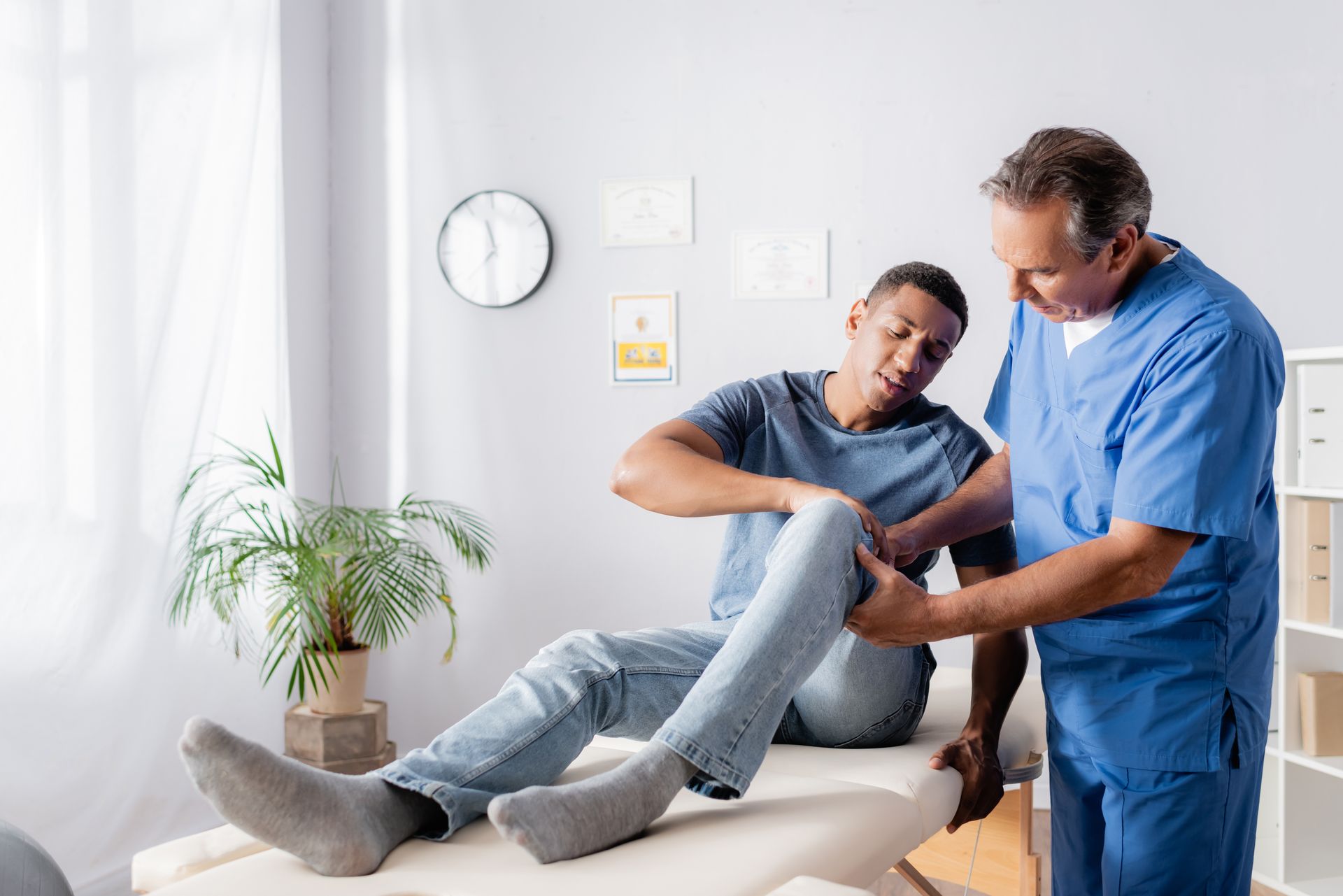 A mature doctor helps and touches the injured knee of a male patient.