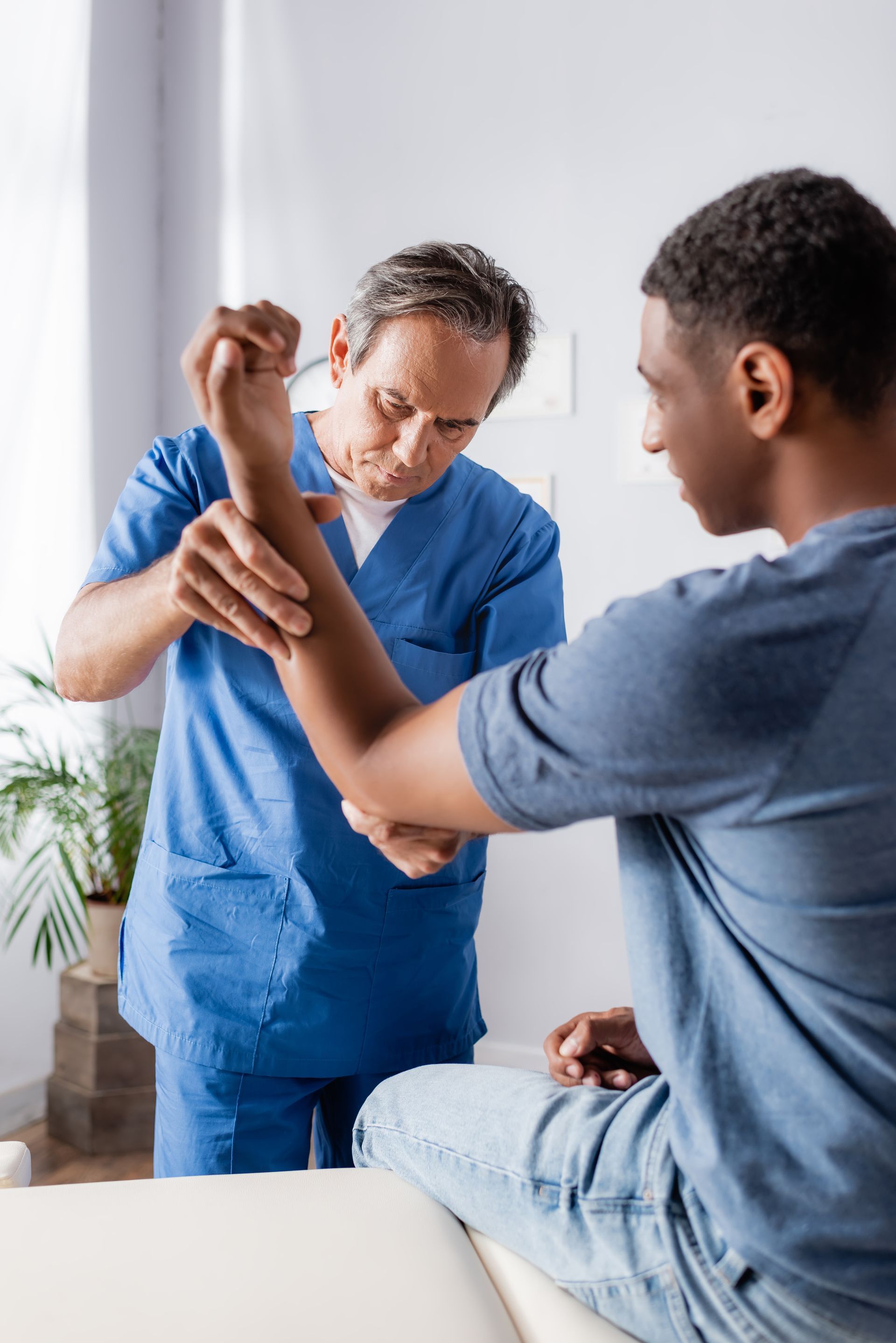 Doctor in blue scrubs examining a young man's injured arm and elbow during a physical therapy exam. Doctor in blue scrubs examining a young man's injured arm and elbow during a physical therapy exam.