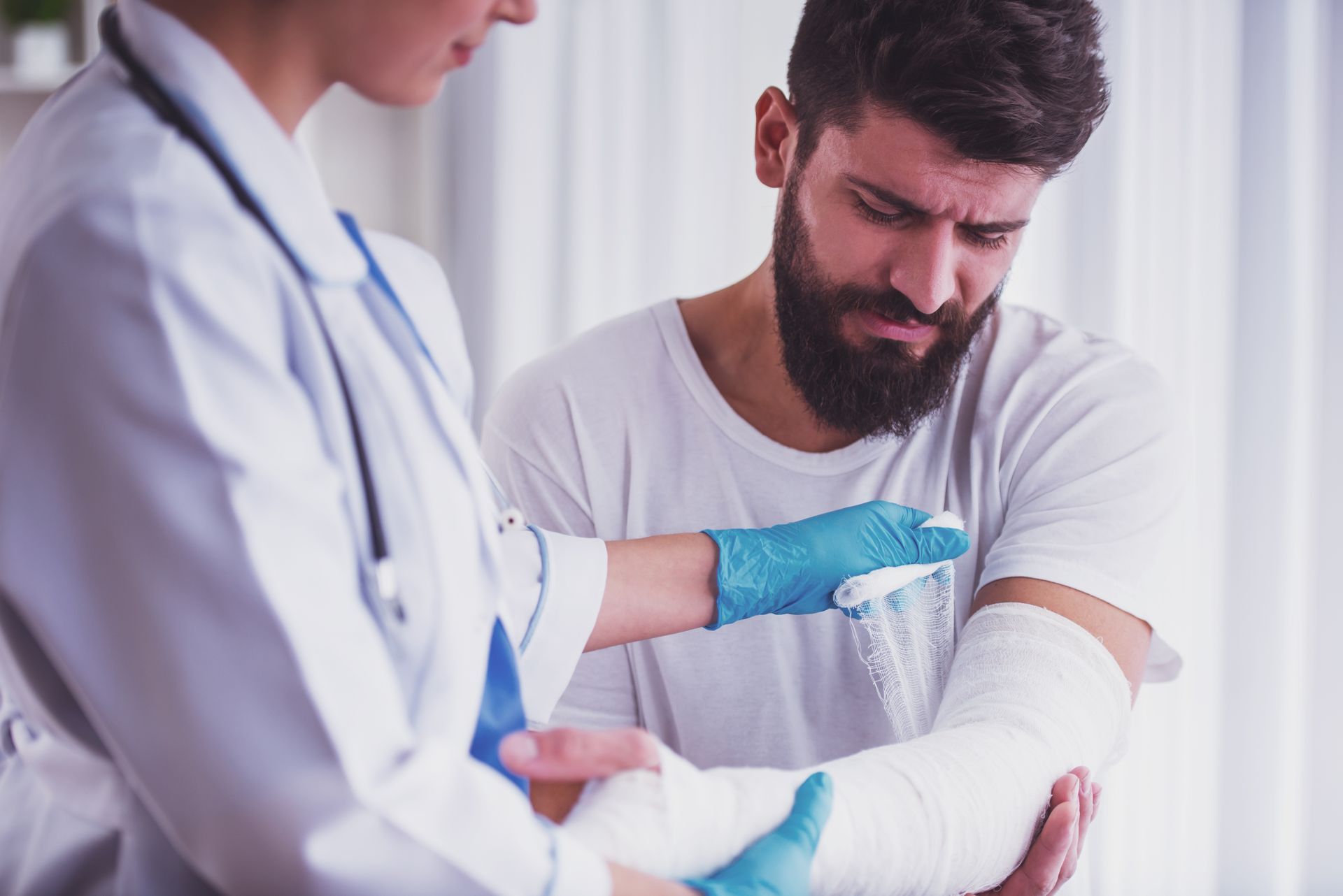 Doctor in white coat and blue gloves wrapping a bandage around a wincing bearded man's injured arm.