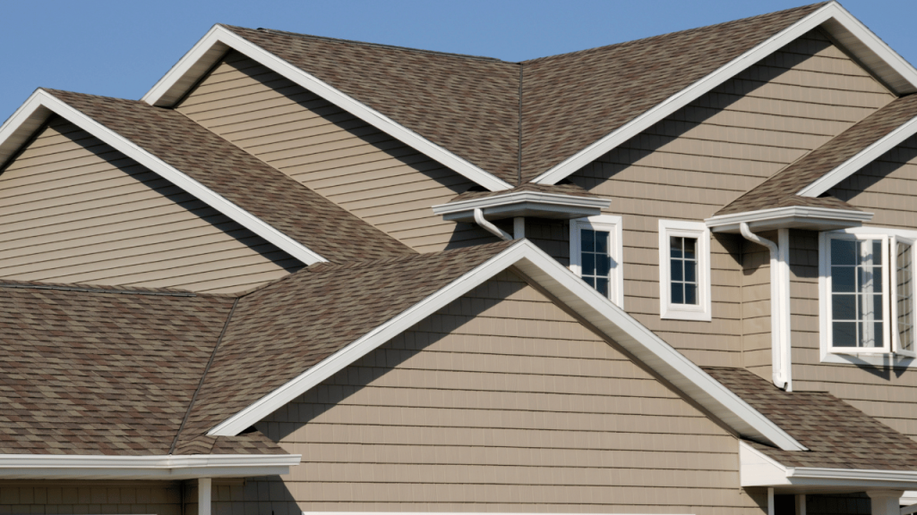 Multi-gabled house with tan siding and brown shingle roof, white trim.