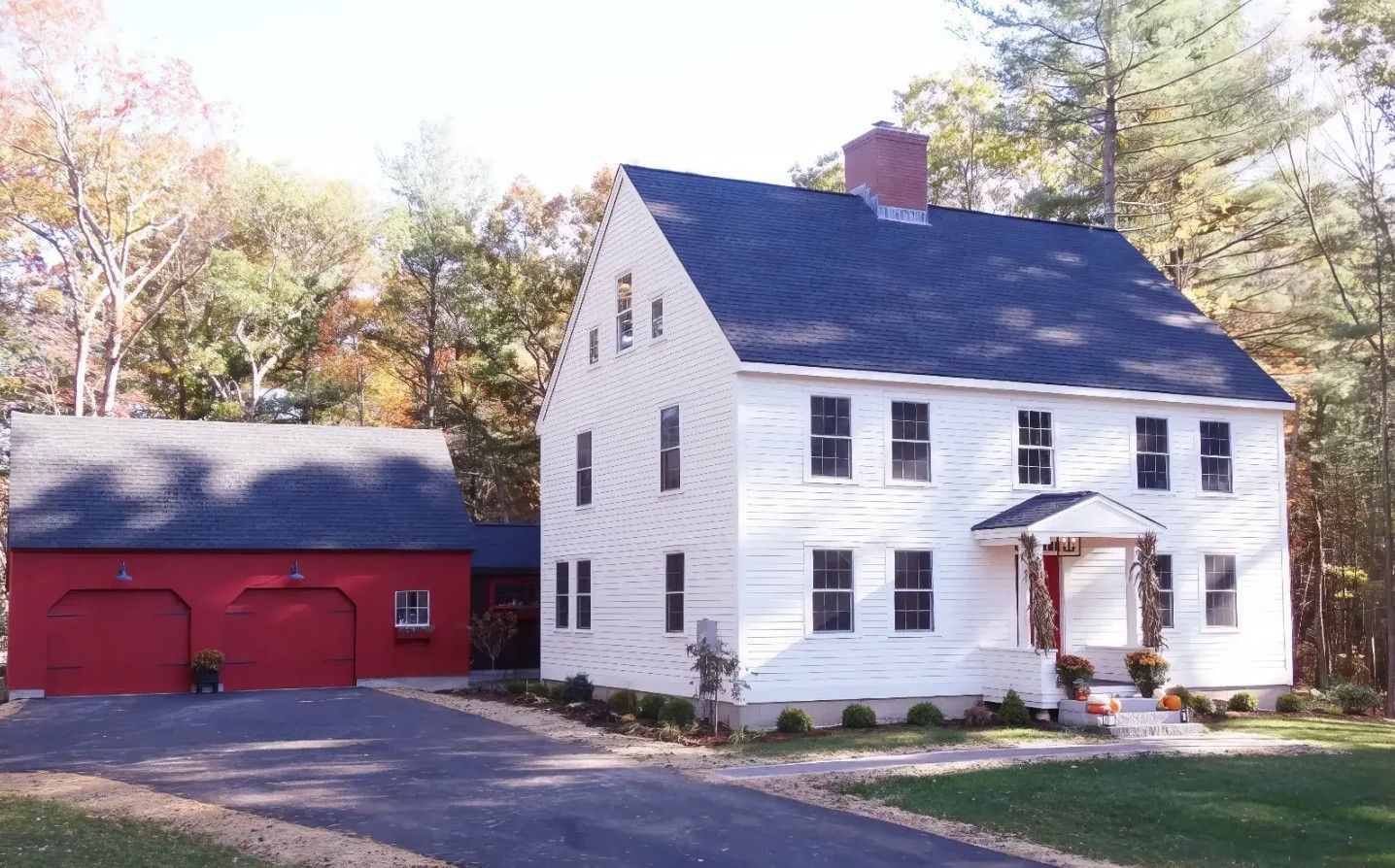 White farmhouse with black roof beside a red barn, surrounded by trees and grass in sunlight
