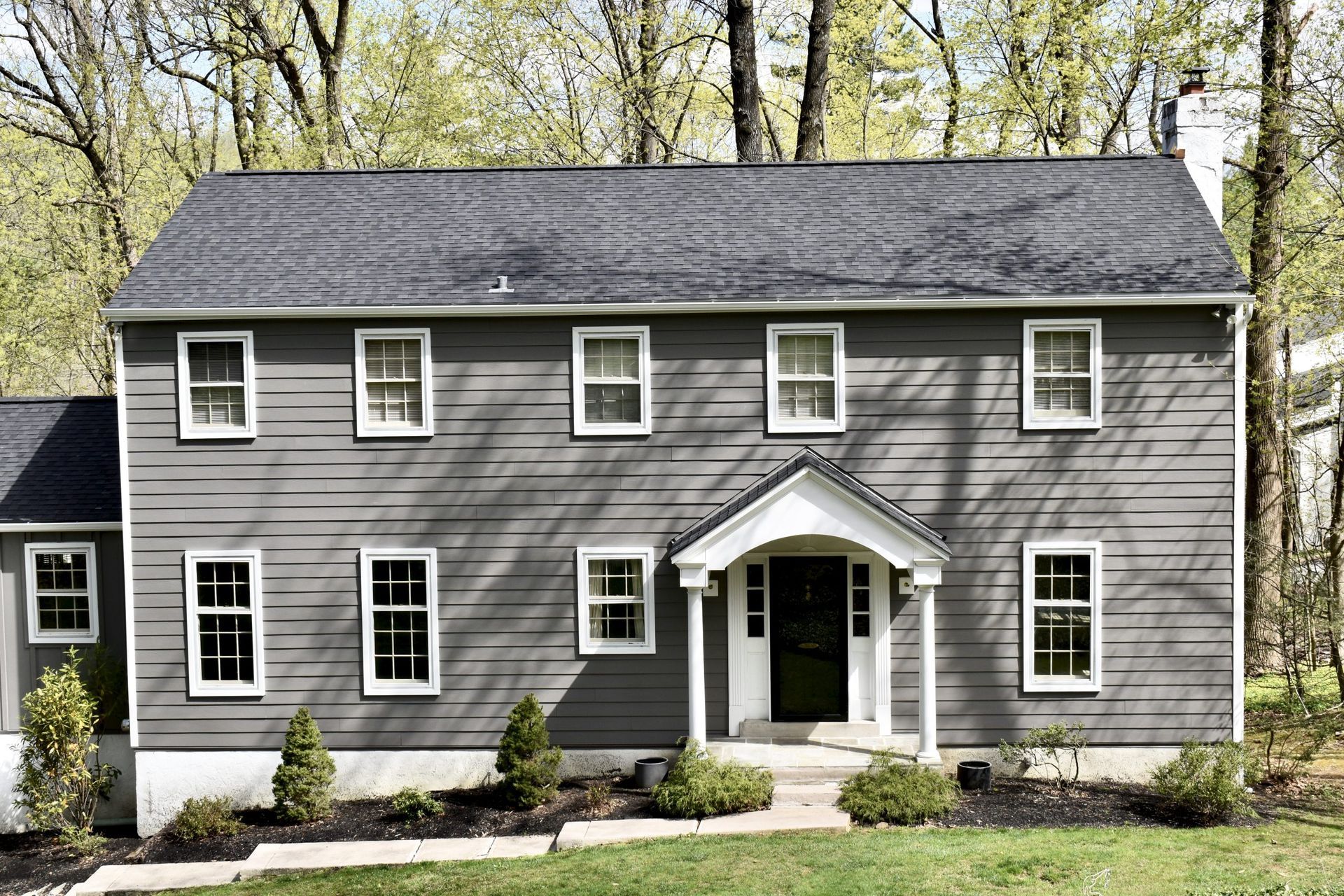 Two-story gray house with white trim and dark roof; front yard with bushes and trees in the background.