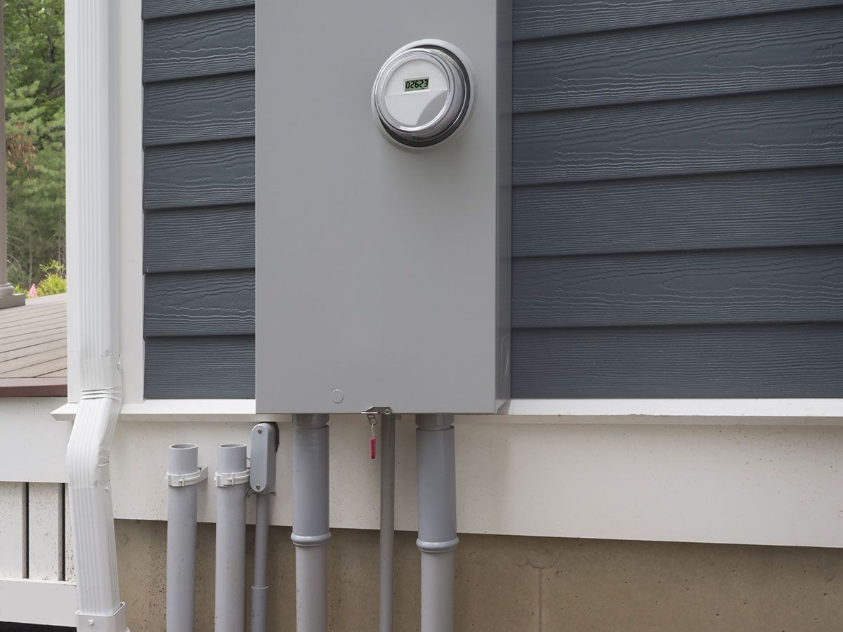 Gray electrical panel on a house exterior, with a meter and conduit pipes.