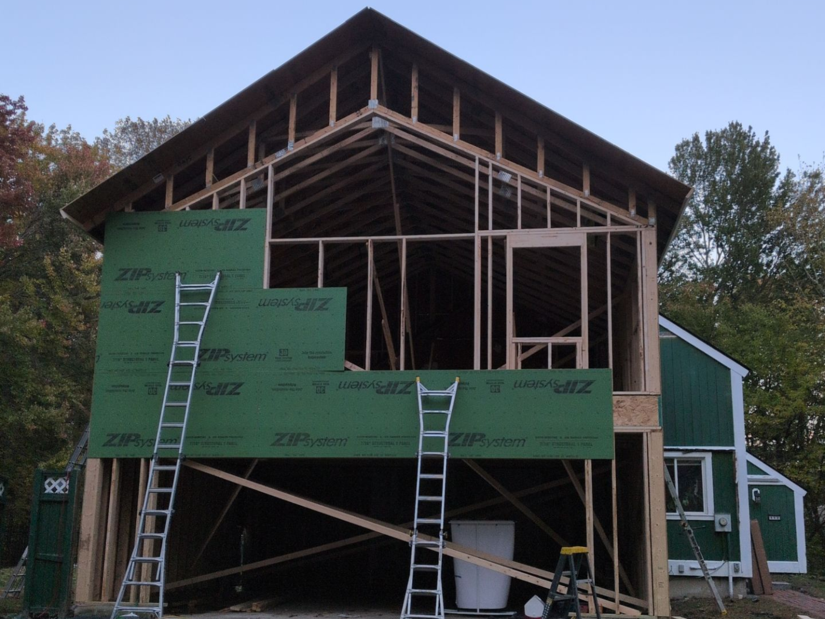 Wooden frame of a building under construction, with green sheathing partially attached and ladders present.
