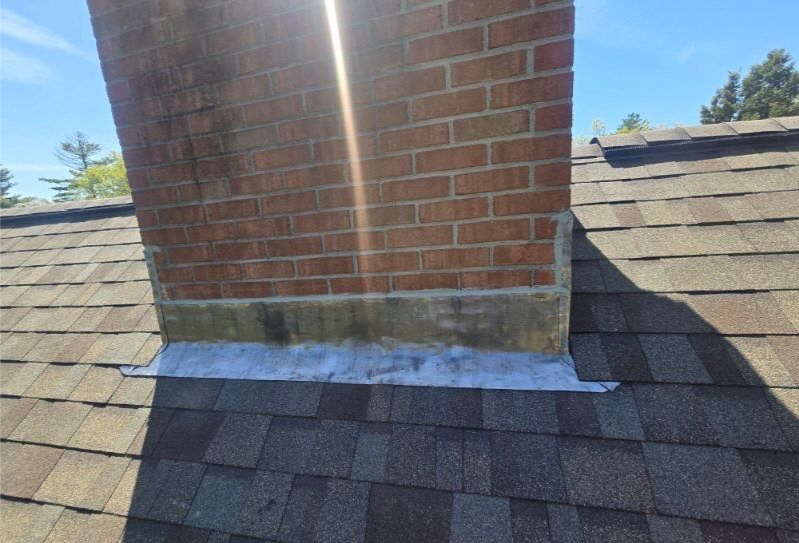 Brick chimney on a shingled roof with flashing, viewed from below on a sunny day.