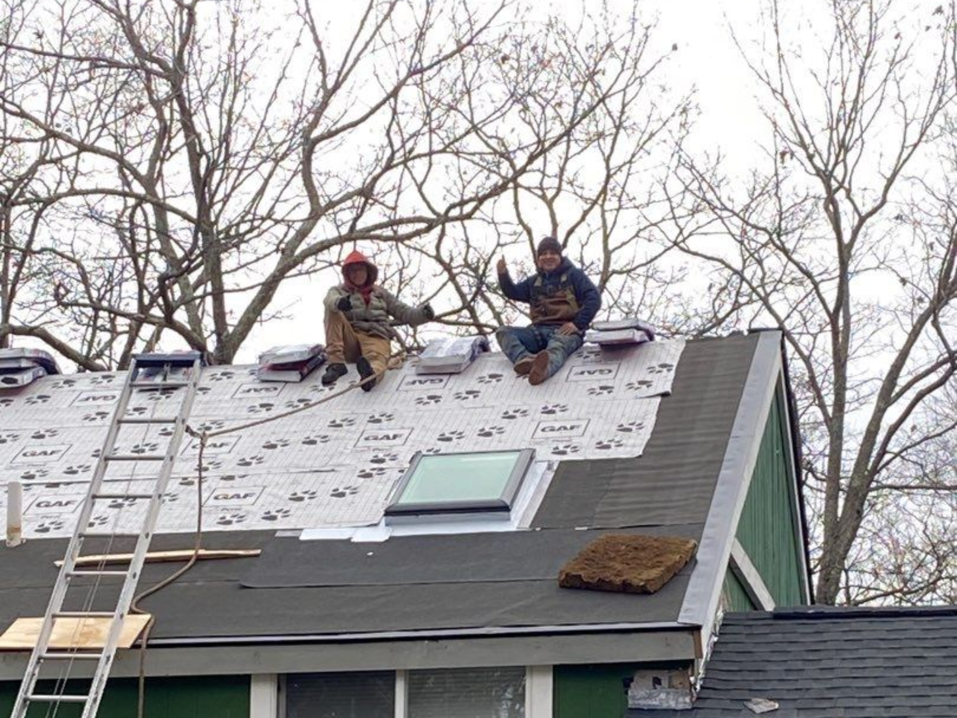 Two workers on a roof, installing shingles, with thumbs up. Green house, clear sky, ladder visible.