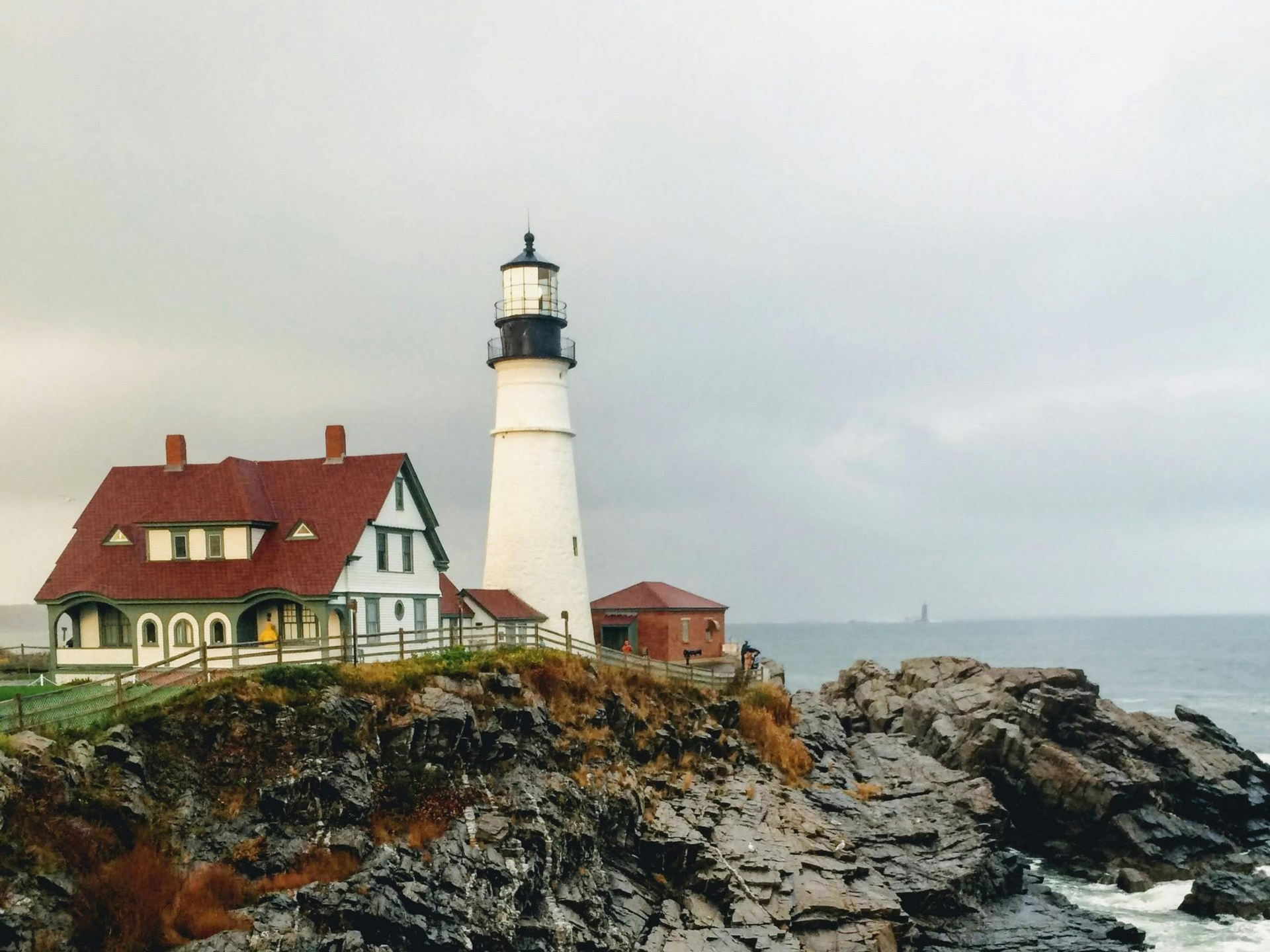 Lighthouse beside a red-roofed house on a rocky coastline under a cloudy sky