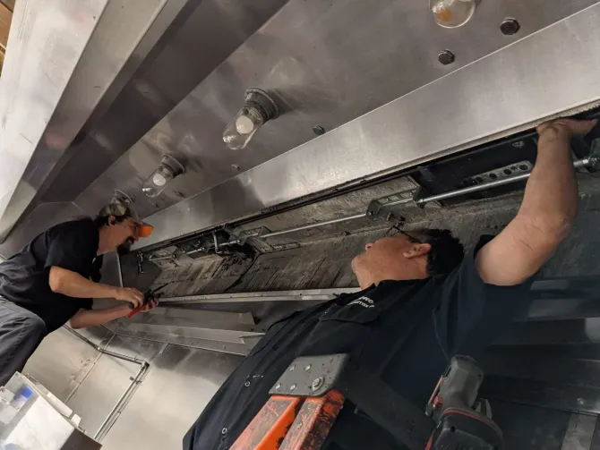 Two technicians working under a metal machine, inspecting wiring and components in an industrial setting