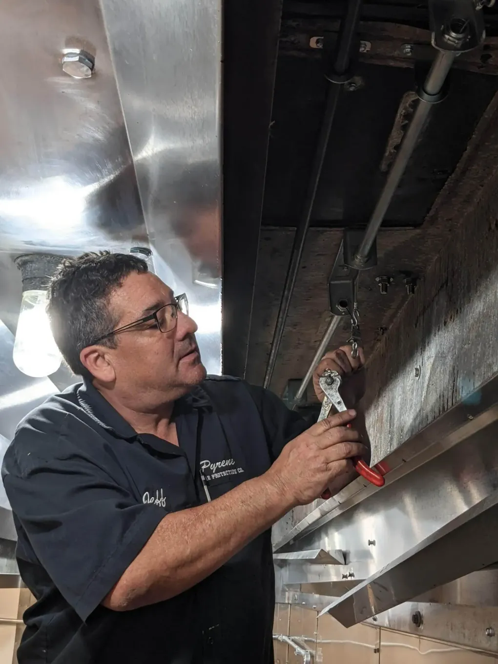 Technician tightening a bolt under metal equipment, working with a wrench in a dim industrial setting