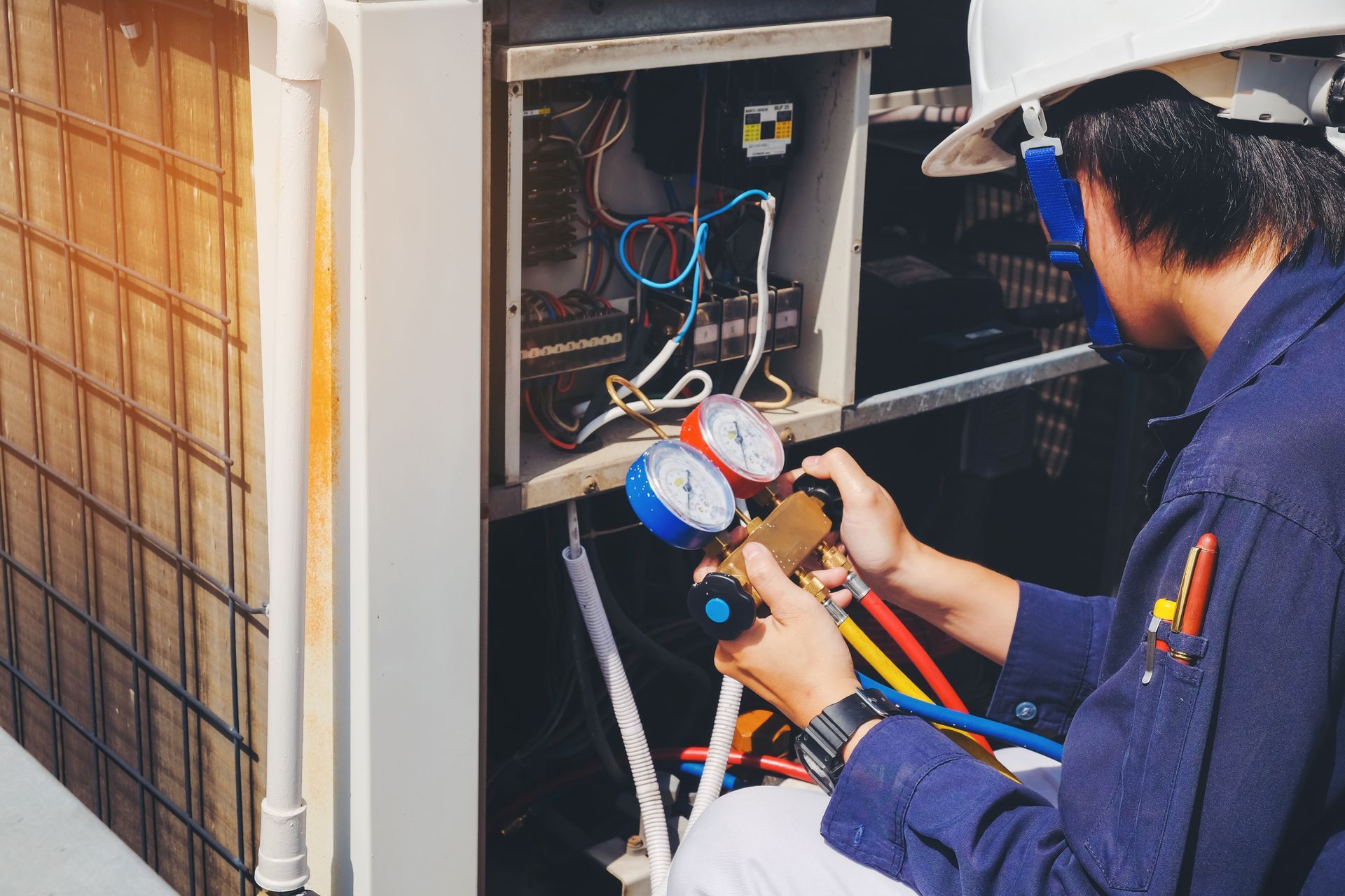 HVAC technician in a blue uniform, wearing a hard hat, working on outdoor AC unit, using gauges. HVAC technician in a blue uniform, wearing a hard hat, working on outdoor AC unit, using gauges.