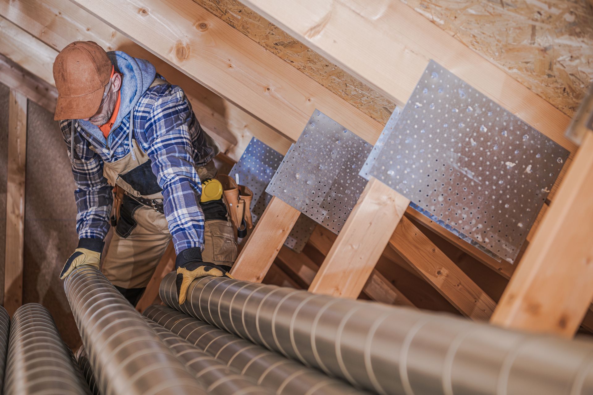 Construction worker in an attic installing HVAC ductwork, wooden beams, metallic tubing, wearing gloves and a hard hat.