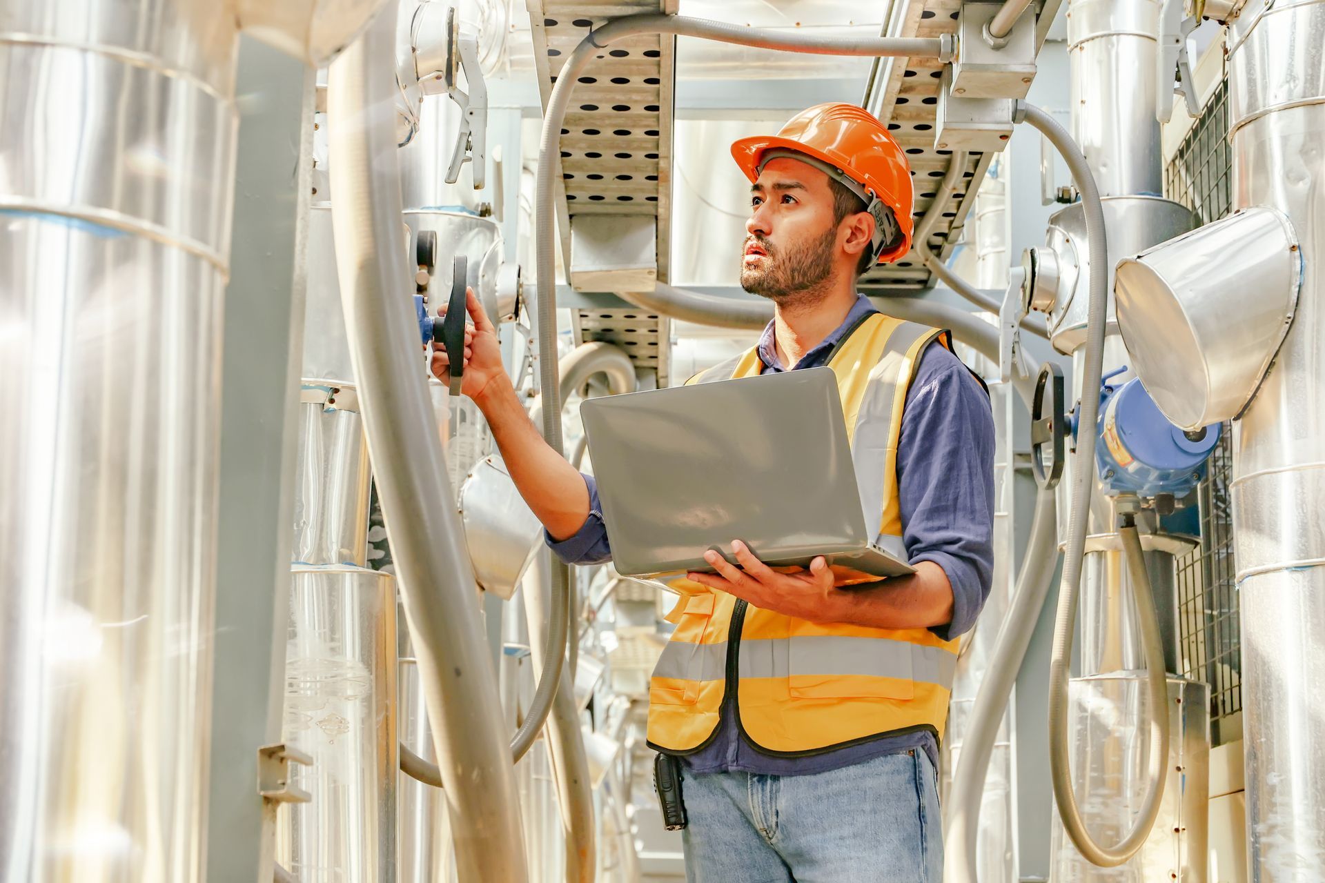 Engineer in hard hat and safety vest, using a laptop in an industrial facility, surrounded by pipes.
