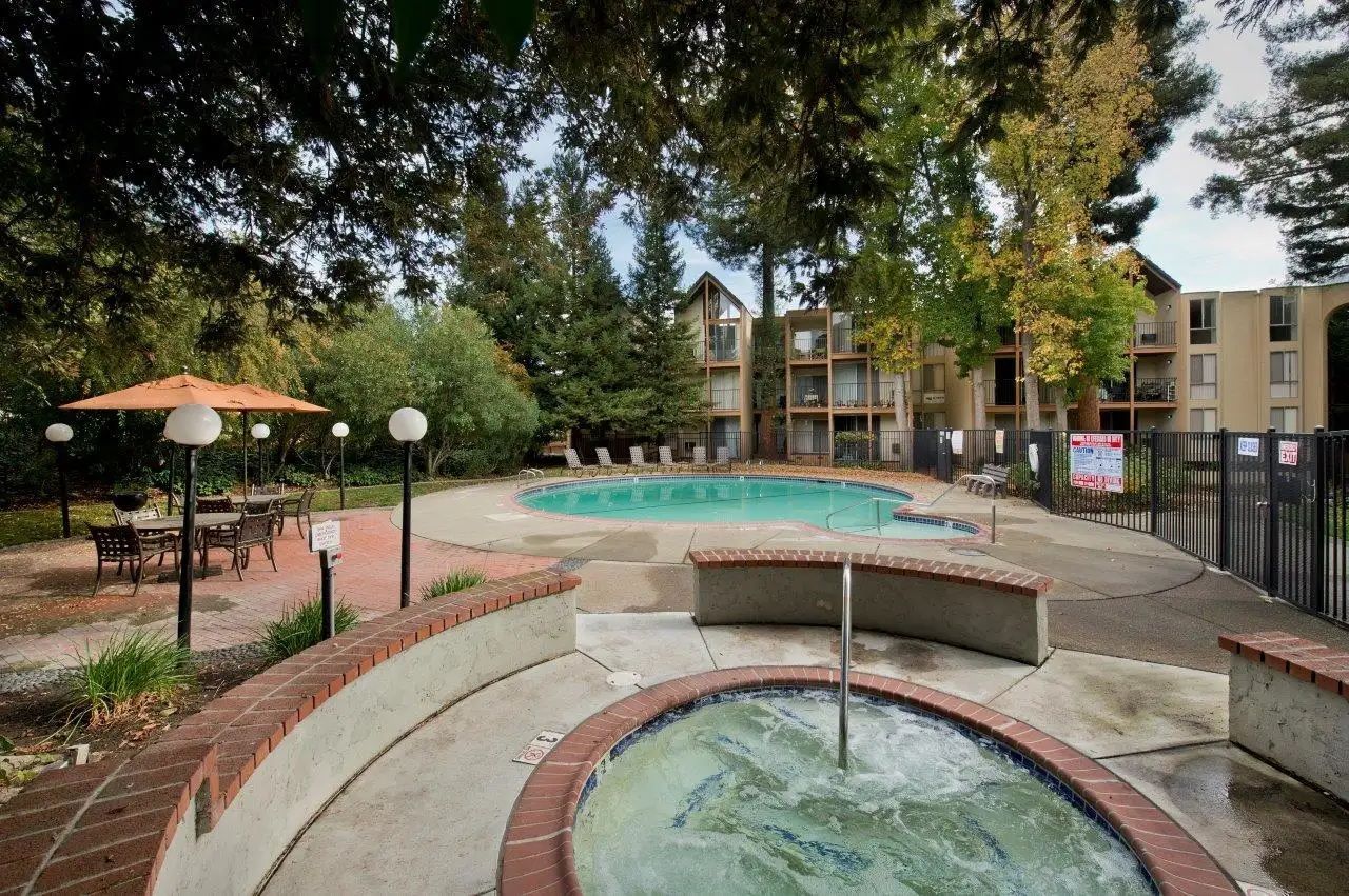 Pool and hot tub area in an apartment complex with seating, surrounded by trees, and buildings in the background.
